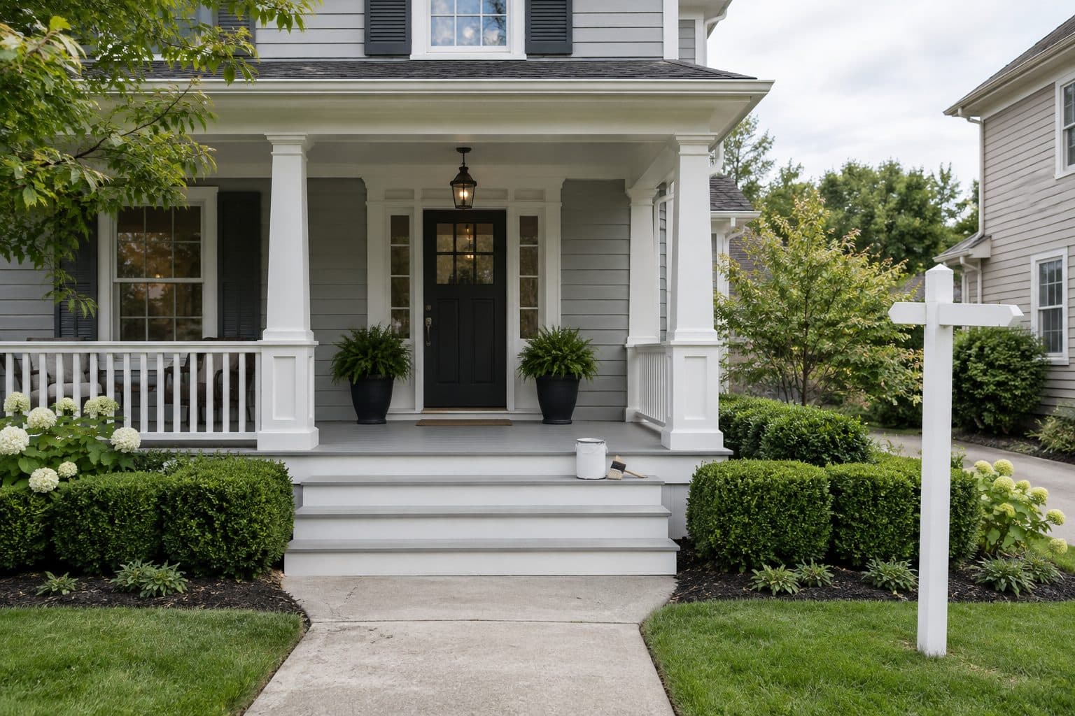 A freshly painted front porch with a paint can on the top step, trimmed hedges, and a bare sign post on the lawn